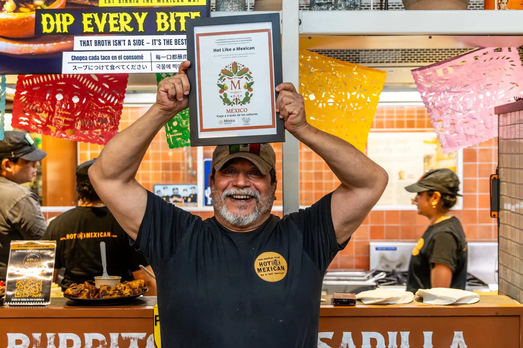 Andrés holds the Sello M certificate above his head in front of the kitchen
